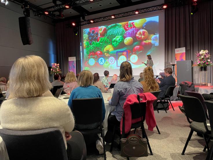 Bilde av publikum med storskjerm i bakgrunnen hvor vi ser fargerike frukter og grønnsaker som illustrerer "fem om dagen" for livskvalitet
