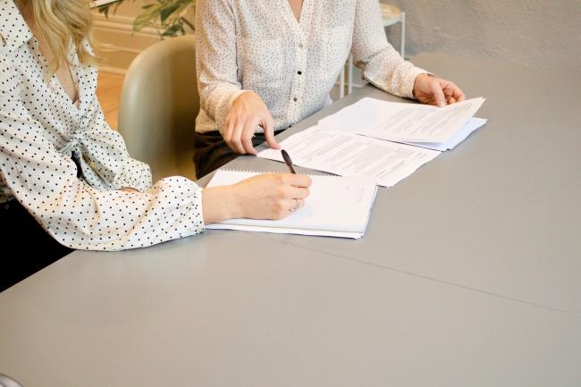 Illustrasjonsbilde - to damer som sitter ved et bord, snakker sammen og noterer på ark.