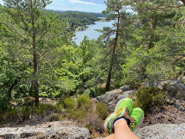 Bilde av bena til en person som sitter på toppen av et fjell med masse grønne trær og utsikt til en sjø