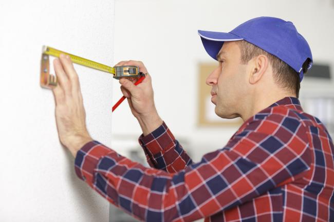 Bilde av en mann med caps som måler noe på en vegg ved hjelp av målebånd
